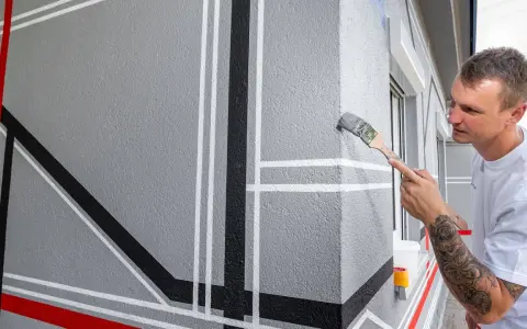 Mann streicht geometrische Muster auf eine graue Fassade. Die Temperatur nachts ist ideal für das Streichen.