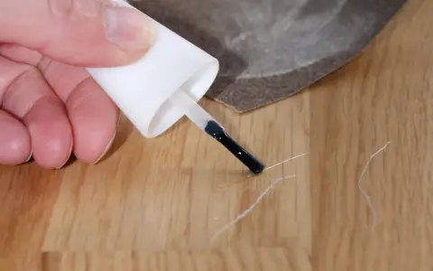 Kleine Kratzer im Parkett reparieren: Ein Finger hält einen Lackstift über eine Holzoberfläche, daneben liegt Schleifpapier.