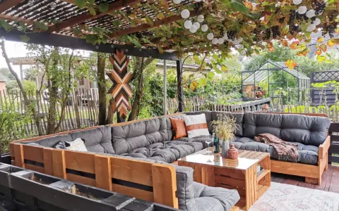 Gemütliche Paletten Lounge Ideen für den Garten. Ein großer Ecksofa aus Paletten mit grauen Polstern und Holztisch. Weinreben und Lichterketten überdachen den Sitzbereich.