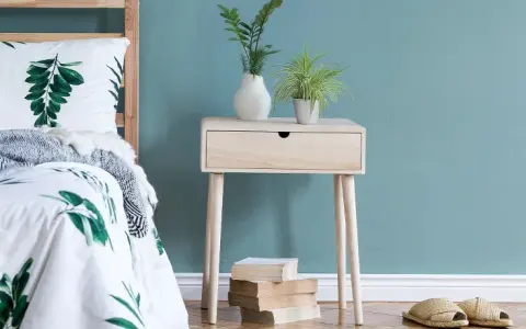 Ein Nachttisch DIY-Projekt aus hellem Holz mit einer Schublade, dekoriert mit Pflanzen und Büchern, neben einem Bett mit Blattmuster.