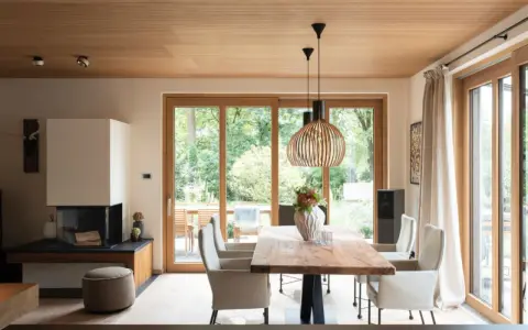 Ein Esszimmer mit Holzboden und Decke, wo sich eiche und buche kombinieren. Ein großer Holztisch steht vor bodentiefen Fenstern.