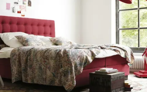 Rotes Boxspringbett mit gemustertem Überwurf und Kissen, ideal für Allergiker. Ein Nachttisch mit Büchern steht daneben.