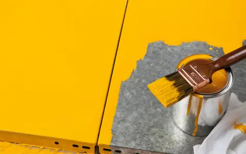 Gelbe Farbe tropft von einem Pinsel, der über eine Dose mit leuchtend gelbem Lack gehalten wird. Ein Teil einer gelben Oberfläche ist sichtbar, während daneben eine graue Fläche mit Farbe bedeckt wird.
