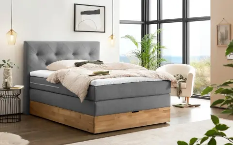 Gemütliches Boxspringbett ohne Lattenrost mit grauem Stoffbezug und Holzsockel. Einladend mit Decken und Kissen dekoriert, bereit für erholsame Nächte.