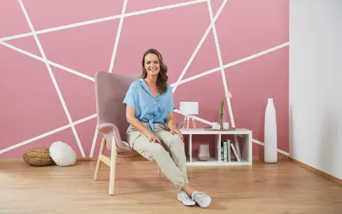 Frau sitzt vor einer rosa Wand, die mit weißen Linien diagonal gestrichen ist.