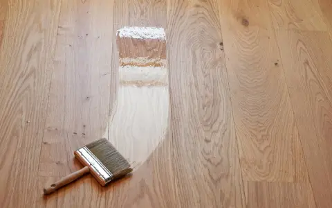 Breite Pinsel mit Holzgriff trägt Lack auf Parkett auf. Der Prozess, das Parkett zu versiegeln, zeigt den Unterschied zwischen unbehandeltem und frisch lackiertem Holz.