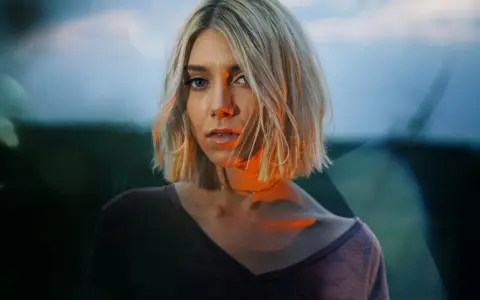 Blonde Frau mit Bob-Schnitt, deren Gesicht von warmem Licht beleuchtet wird, das sich mit dem blauen Abendhimmel mischt.