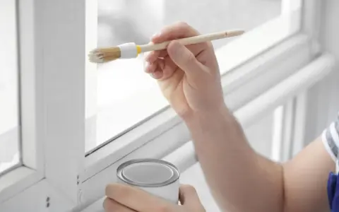 Hand streicht mit Pinsel und weißer Farbe einen weißen Kunststoffrahmen. Dose mit Farbe steht bereit.