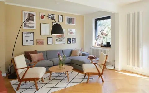 Gemütliches Wohnzimmer mit grauem Sofa, zwei Sesseln und vielen Bildern an der gelben Wand. Perfekt, um die Wand abzukleben und neu zu gestalten.