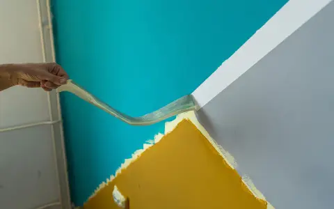 Hand zieht Klebeband ab, um eine scharfe Kante beim Wand malen zu schaffen. Türkis trifft auf Grau und Ockergelb.