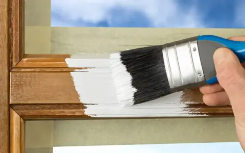 Hand streicht mit weißer Farbe über einen hölzernen Rahmen, um ihn aufzufrischen. Der Spiegelrahmen wird sorgfältig mit Klebeband geschützt.