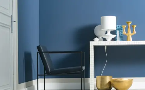 Ein Sessel vor einer Wand in moderner Wandfarbe. Auf einem Regal stehen eine Lampe, eine Vase und ein goldener Kerzenständer.