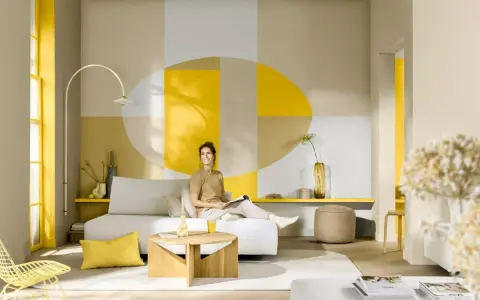 Eine Frau sitzt auf einem Sofa in einem hellen Wohnzimmer. Die Wand hinter ihr ist mit geometrischen Formen in Gelb, Beige und Grau gestaltet. Die Frage, welche Farbe passt zu Gelb, wird hier mit warmen Erdtönen und neutralen Farben beantwortet.
