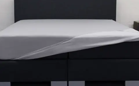 Ein graues Spannbettlaken wird über ein Boxspringbett gezogen. So beziehe ich ein Boxspringbett – einfach und schnell.