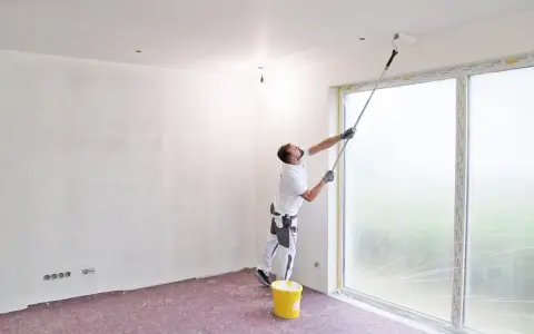 Maler streicht Decke und Wand mit langem Teleskoproller, um das Streichen ohne Abkleben zu ermöglichen. Gelber Farbeimer steht bereit.