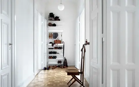 Schmaler Flur mit weißer Wandfarbe, Holzboden und einem Regal voller Schuhe und Hüte. Ein Stuhl steht bereit.