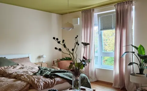 Gemütliches Schlafzimmer mit Bett in Khaki-Farbe, grüner Decke und rosa Vorhängen. Ein Blumenstrauß steht auf einem Holztisch.