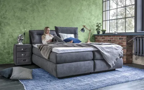 Frau entspannt auf einem Boxspringbett mit grauem Stoffbezug. Das Bett bietet hohen Komfort, was ein Boxspringbett ausmacht.