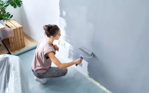 Frau streicht Wand grau. Nützliche **Wand streichen Tipps** für ein gleichmäßiges Ergebnis. Möbeltisch mit Folie abgedeckt.