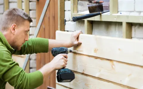 Mann renoviert Holzfassade, schraubt neue Bretter an. Werkzeug liegt bereit.