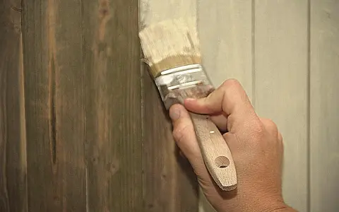 Hand streicht mit Pinsel über dunkles Holz, um es aufzuhellen. Ein Teil der Wand ist bereits heller gestrichen.