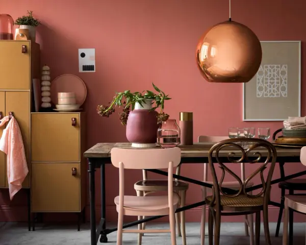 Ein Esszimmer mit altrosa Wänden. Ein kupferfarbener Lampenschirm und ein Holzmöbelstück mit gelben Türen. Welche Farbe zu altrosa? Zarte Rosatöne und Erdtöne passen gut.