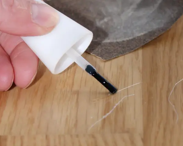 Kleine Kratzer im Parkett reparieren: Ein Finger hält einen Lackstift über eine Holzoberfläche, daneben liegt Schleifpapier.