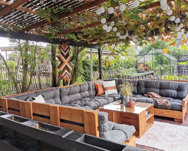 Gemütliche Paletten Lounge Ideen für den Garten. Ein großer Ecksofa aus Paletten mit grauen Polstern und Holztisch. Weinreben und Lichterketten überdachen den Sitzbereich.