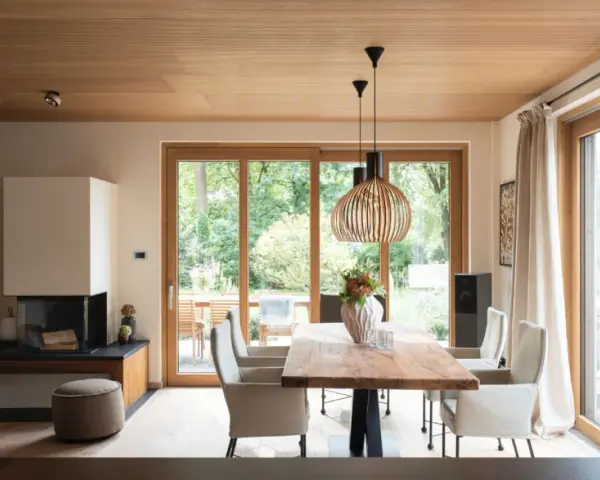 Ein Esszimmer mit Holzboden und Decke, wo sich eiche und buche kombinieren. Ein großer Holztisch steht vor bodentiefen Fenstern.