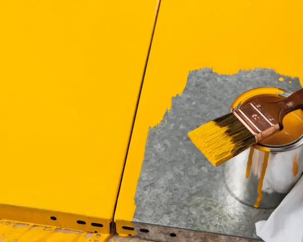 Gelbe Farbe tropft von einem Pinsel, der über eine Dose mit leuchtend gelbem Lack gehalten wird. Ein Teil einer gelben Oberfläche ist sichtbar, während daneben eine graue Fläche mit Farbe bedeckt wird.