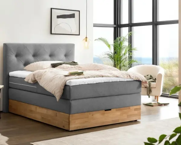 Gemütliches Boxspringbett ohne Lattenrost mit grauem Stoffbezug und Holzsockel. Einladend mit Decken und Kissen dekoriert, bereit für erholsame Nächte.