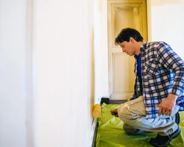 Mann kniet auf grüner Folie und trägt mit einer Rolle eine neue Farbe auf, um alte Latexfarbe zu entfernen und die Wand zu renovieren.