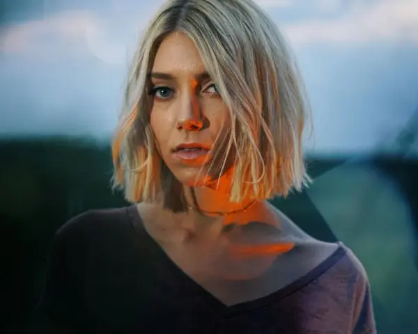 Blonde Frau mit Bob-Schnitt, deren Gesicht von warmem Licht beleuchtet wird, das sich mit dem blauen Abendhimmel mischt.