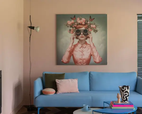 Ein blauer Sofa mit Kissen vor einer Wand mit einem surrealen Gemälde, das eine Frau mit Blumen und Schmetterlingen auf dem Kopf zeigt. Eine kreative Wandgestaltung, die zum Träumen einlädt.