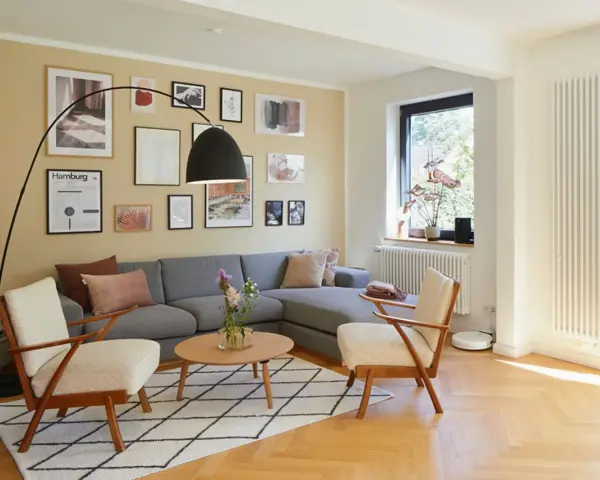 Gemütliches Wohnzimmer mit grauem Sofa, zwei Sesseln und vielen Bildern an der gelben Wand. Perfekt, um die Wand abzukleben und neu zu gestalten.
