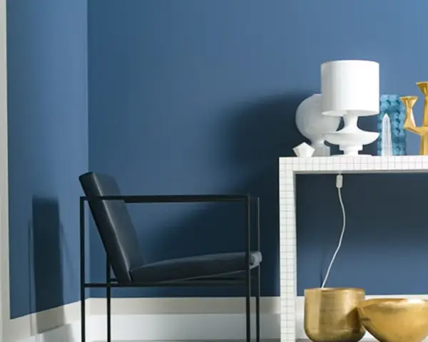 Ein Sessel vor einer Wand in moderner Wandfarbe. Auf einem Regal stehen eine Lampe, eine Vase und ein goldener Kerzenständer.