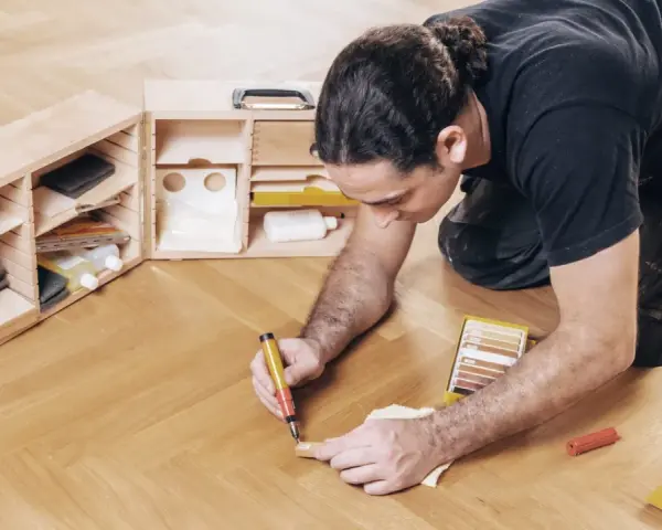 Mann repariert den Holzboden mit einem Reparaturstift und einer Farbkarte. Werkzeugkasten mit Zubehör daneben.