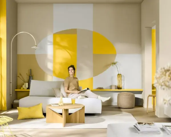 Eine Frau sitzt auf einem Sofa in einem hellen Wohnzimmer. Die Wand hinter ihr ist mit geometrischen Formen in Gelb, Beige und Grau gestaltet. Die Frage, welche Farbe passt zu Gelb, wird hier mit warmen Erdtönen und neutralen Farben beantwortet.
