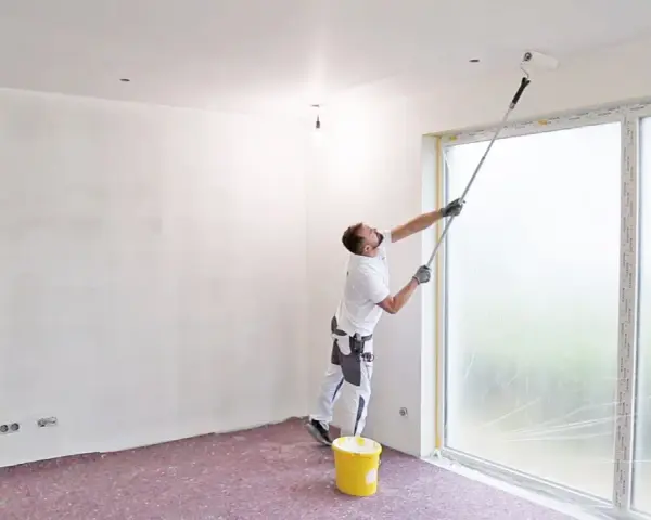 Maler streicht Decke und Wand mit langem Teleskoproller, um das Streichen ohne Abkleben zu ermöglichen. Gelber Farbeimer steht bereit.