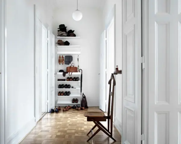 Schmaler Flur mit weißer Wandfarbe, Holzboden und einem Regal voller Schuhe und Hüte. Ein Stuhl steht bereit.