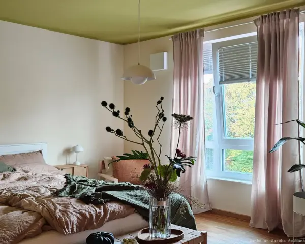 Gemütliches Schlafzimmer mit Bett in Khaki-Farbe, grüner Decke und rosa Vorhängen. Ein Blumenstrauß steht auf einem Holztisch.