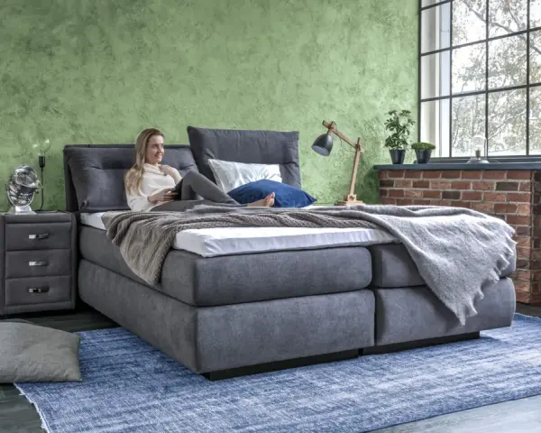 Frau entspannt auf einem Boxspringbett mit grauem Stoffbezug. Das Bett bietet hohen Komfort, was ein Boxspringbett ausmacht.