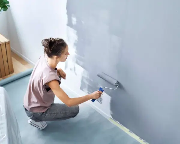 Frau streicht Wand grau. Nützliche **Wand streichen Tipps** für ein gleichmäßiges Ergebnis. Möbeltisch mit Folie abgedeckt.
