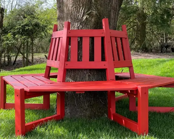 Frisch gestrichene rote Gartenbank um einen Baum im Grünen.