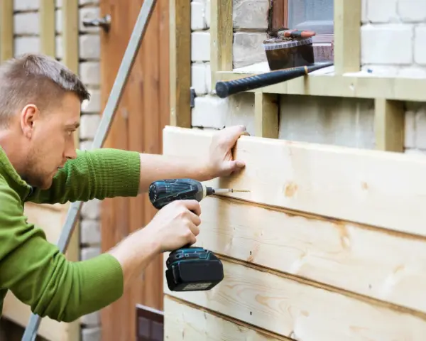 Mann renoviert Holzfassade, schraubt neue Bretter an. Werkzeug liegt bereit.