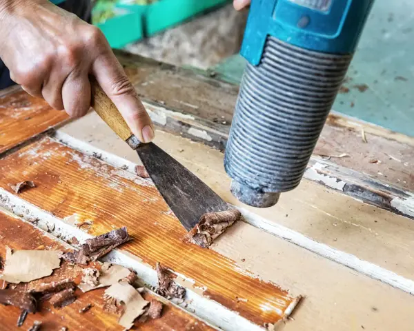 Farbe von Holz entfernen ohne Schleifen: Heißluftföhn und Spachtel lösen alte Lackschichten von einer Holztür.