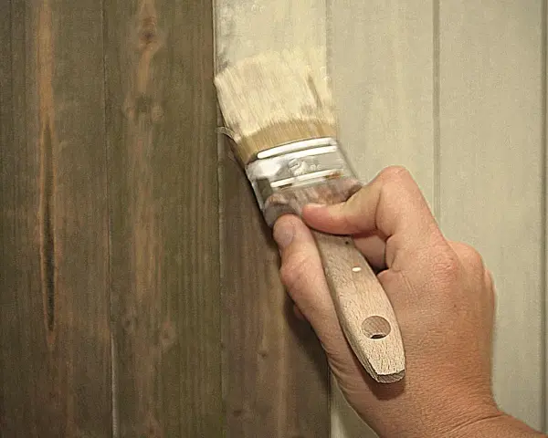 Hand streicht mit Pinsel über dunkles Holz, um es aufzuhellen. Ein Teil der Wand ist bereits heller gestrichen.