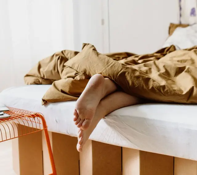 Füße ragen aus einem gemütlichen, braunen Bett hervor. Das Bett ist 210 cm lang und lädt zum Entspannen ein.