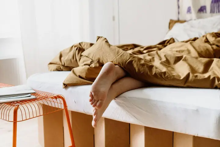 Füße ragen aus einem gemütlichen, braunen Bett hervor. Das Bett ist 210 cm lang und lädt zum Entspannen ein.