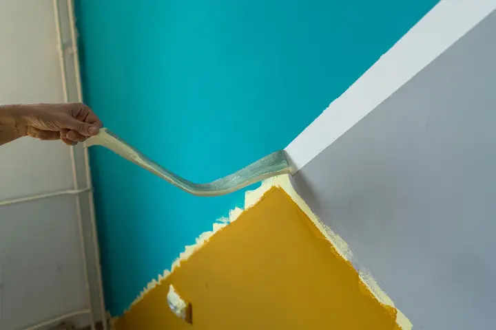 Hand zieht Klebeband ab, um eine scharfe Kante beim Wand malen zu schaffen. Türkis trifft auf Grau und Ockergelb.